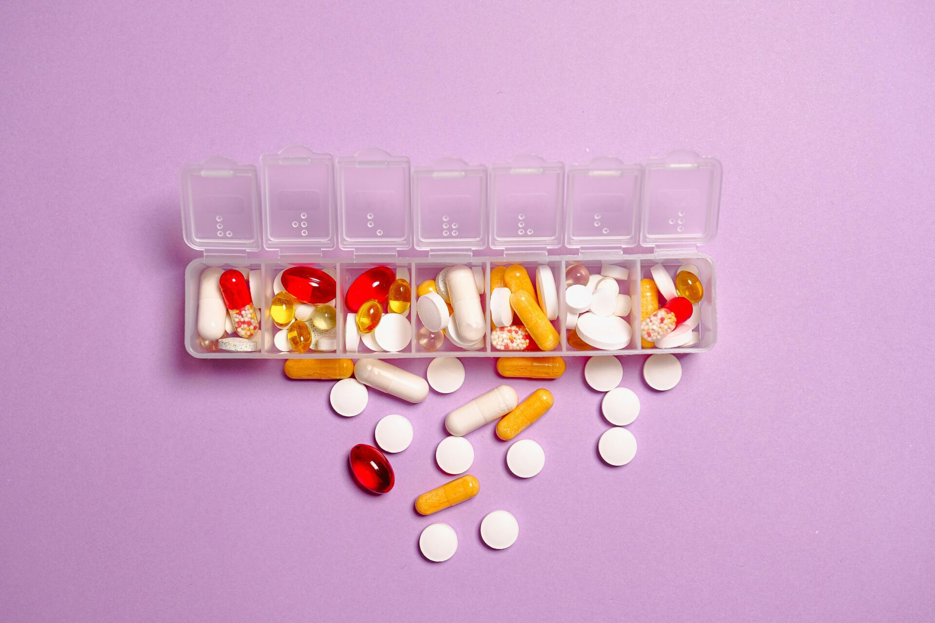 A top-down view of a weekly pill organizer filled with various colourful capsules and tablets on a purple background